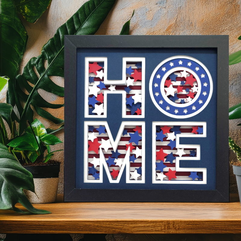 Framed decorative sign with 'HOME' in American flag pattern on a wooden surface with plants.