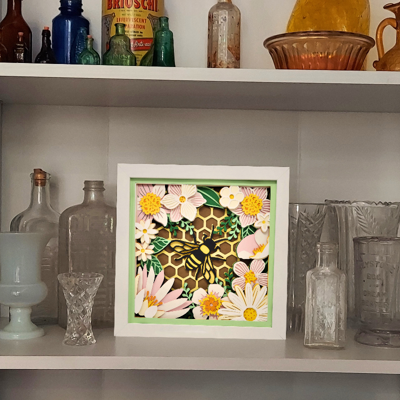 Framed artwork with a bee and floral design on a shelf with vintage bottles and glassware.