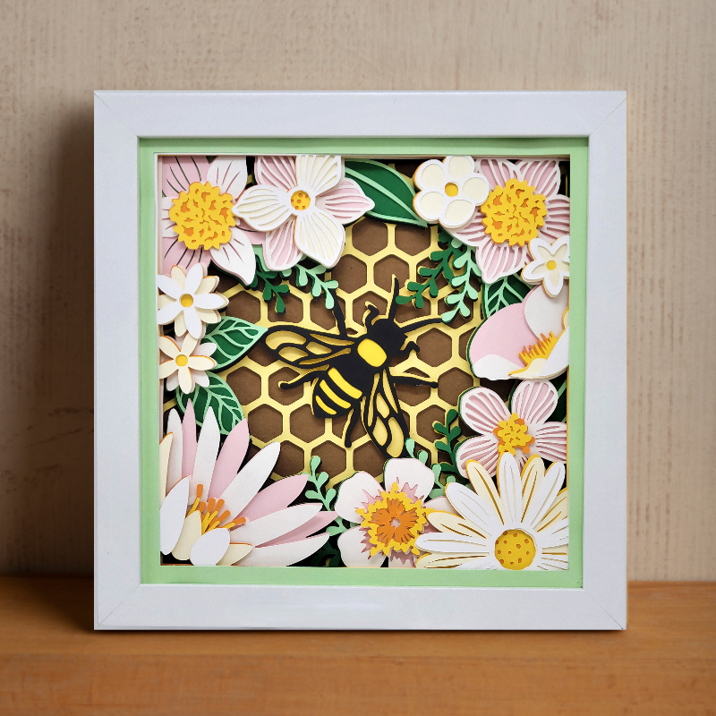 Decorative wall art with a bee and flowers in a white frame.