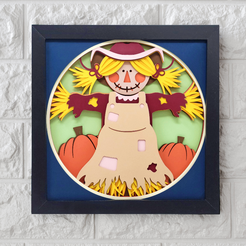 Framed paper cutout of a scarecrow with pumpkins on a white brick wall background