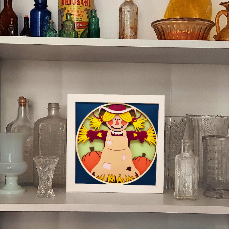 Framed artwork of a scarecrow with pumpkins on a shelf with decorative bottles and glasses.