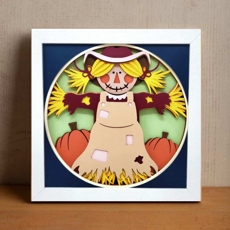 Framed paper cutout of a scarecrow with pumpkins on a wooden surface.