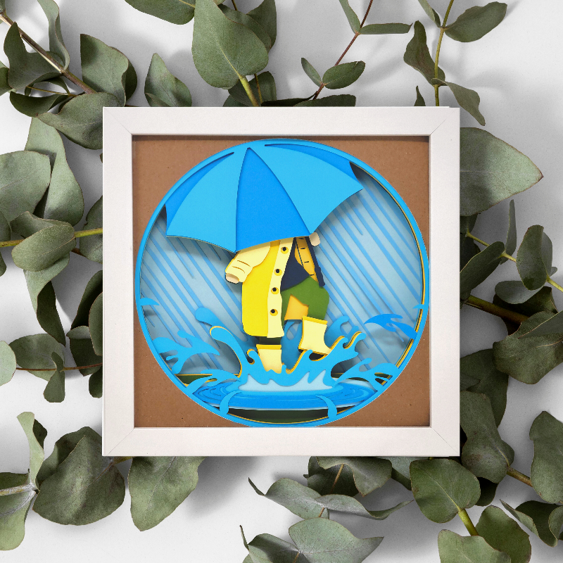 Framed paper art of a person with a blue umbrella and yellow raincoat on a brown background, surrounded by green leaves.