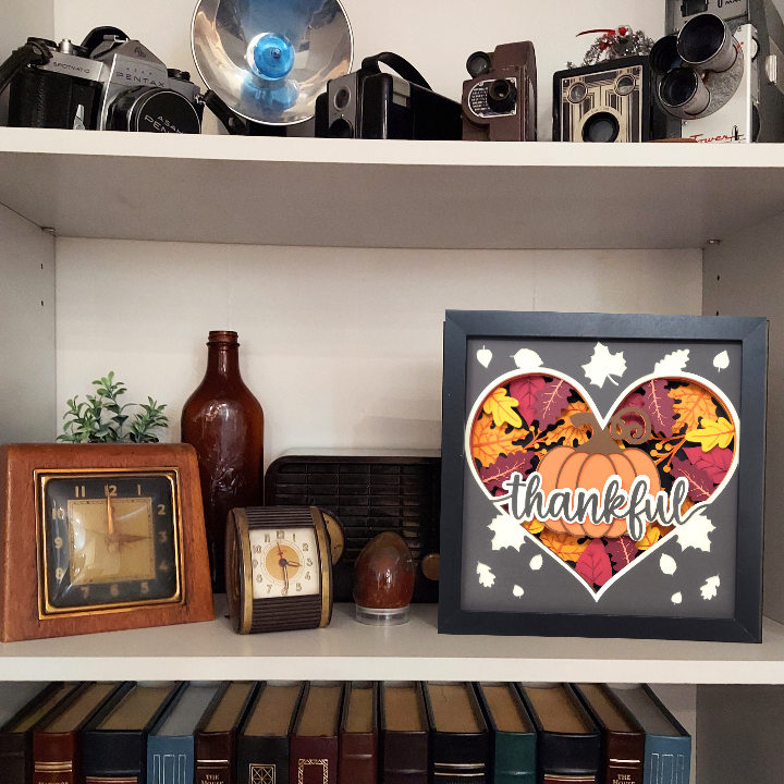 Decorative shelf with vintage cameras, bottles, clocks, and a 'thankful' heart-shaped frame.