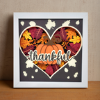 Framed artwork with a pumpkin and leaves inside a heart shape, featuring the word 'thankful'.