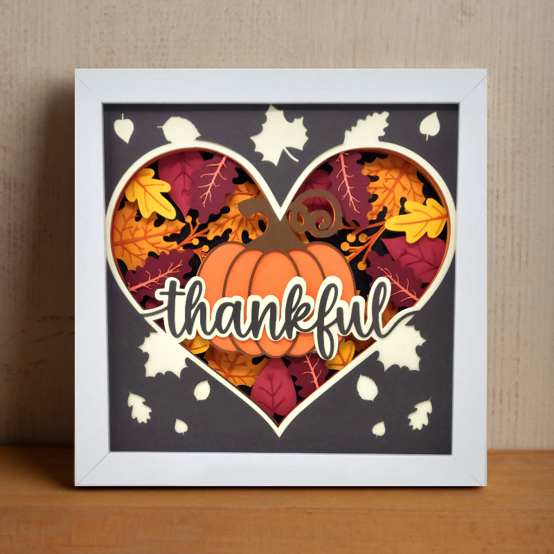 Framed artwork with a pumpkin and leaves inside a heart shape, featuring the word 'thankful'.