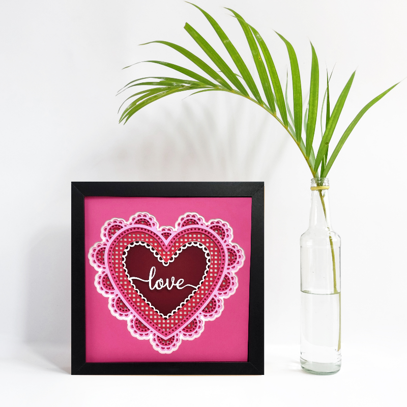 Heart-shaped artwork with 'love' text on a pink background next to a clear vase with greenery.
