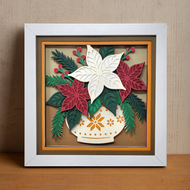 Framed paper art of poinsettias and leaves on a wooden surface with a beige wall background