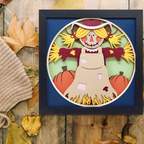 Framed paper cutout of a scarecrow with pumpkins on a wooden surface with autumn leaves.