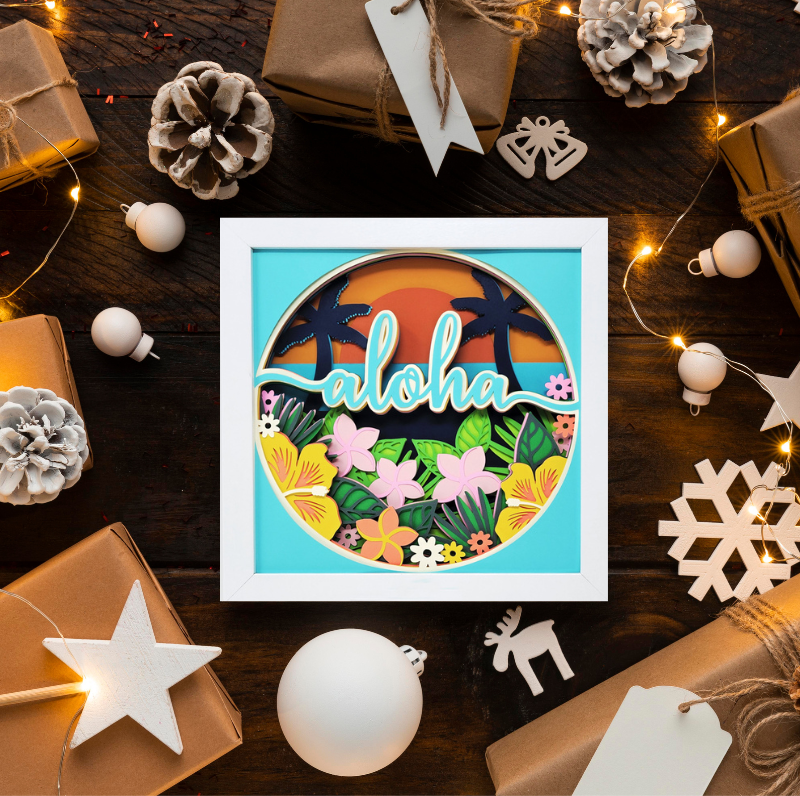 Framed 'aloha' sign with tropical design on a wooden surface with Christmas decorations.