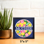 Framed artwork with floral design and 'MOTHER' text on a wooden surface with a plant.