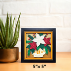 Decorative wall art with floral design in a black frame on a wooden surface next to a plant.