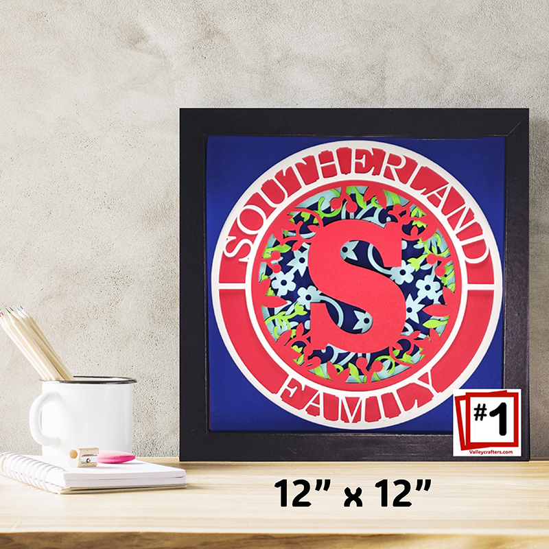 Framed artwork with floral design and letter 'S' on a blue background, placed on a desk.