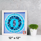 Framed decorative item with blue and white design on a gray wall with a plant.