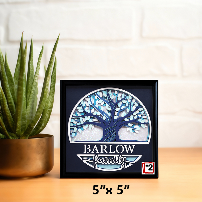 Framed artwork with a tree design and 'Barlow Family' text on a wooden surface with a plant.