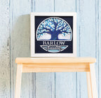 Framed artwork with a tree design and 'Barlow Family' text on a wooden stool against a light blue wooden panel background.