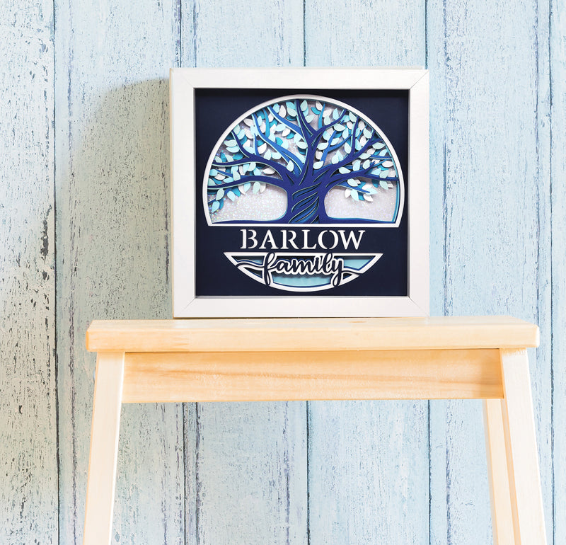Framed artwork with a tree design and 'Barlow Family' text on a wooden stool against a light blue wooden panel background.