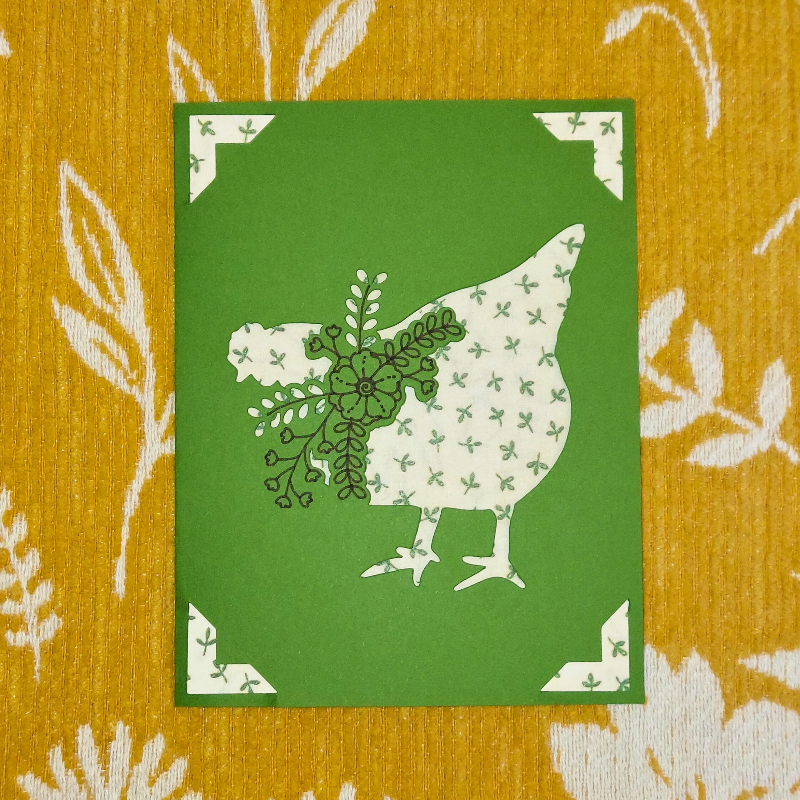 Card with a chicken silhouette and floral design on a green background, placed on a patterned surface.