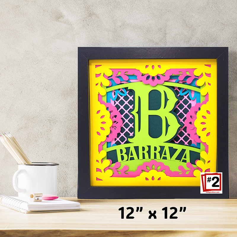 Framed decorative wall art with 'BARRAZA' on a wooden surface with stationery items.