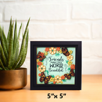 Framed artwork with butterflies and flowers on a wooden surface with a plant in the background