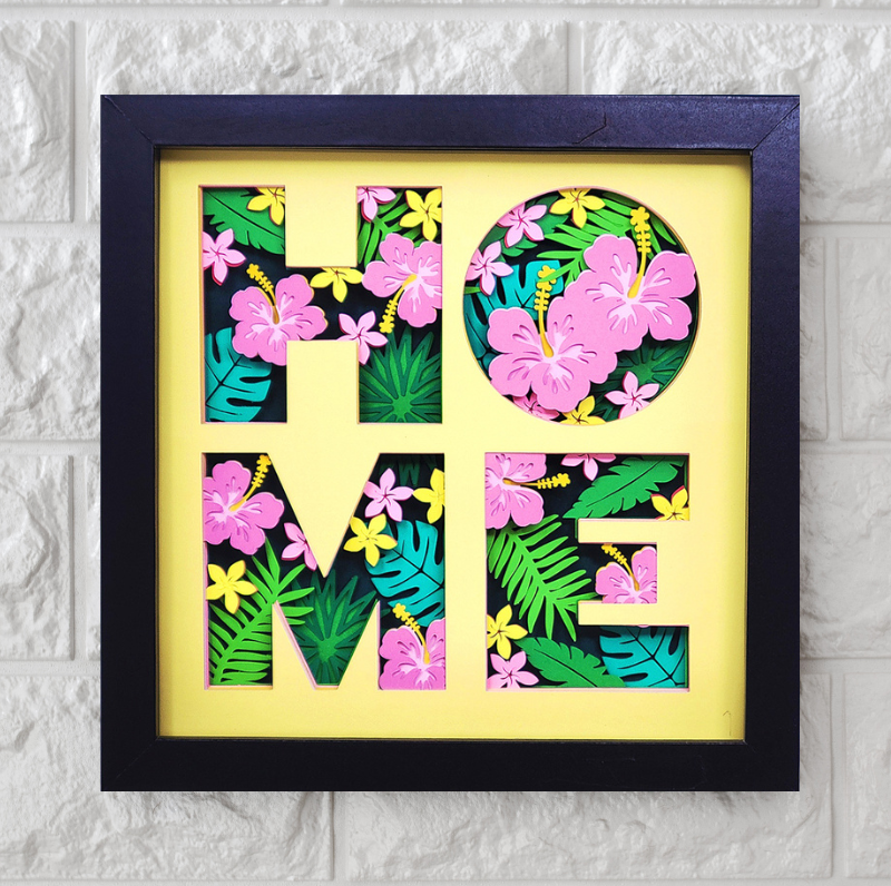 Framed decorative wall art with 'HOME' letters featuring tropical leaves and flowers on a white brick background.
