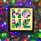 Decorative wall art with 'HOME' letters surrounded by floral patterns on a wooden background with string lights.