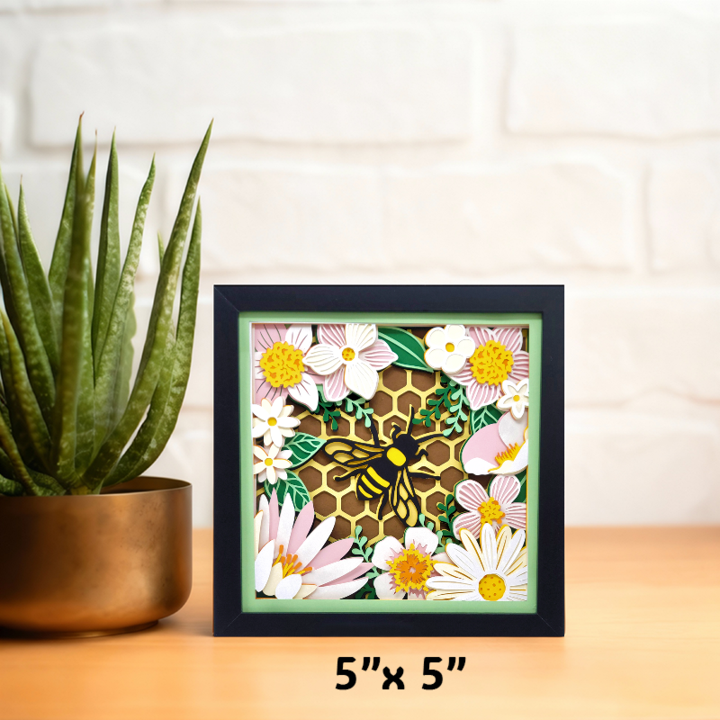 Framed artwork with a bee and floral design on a wooden surface with a plant.