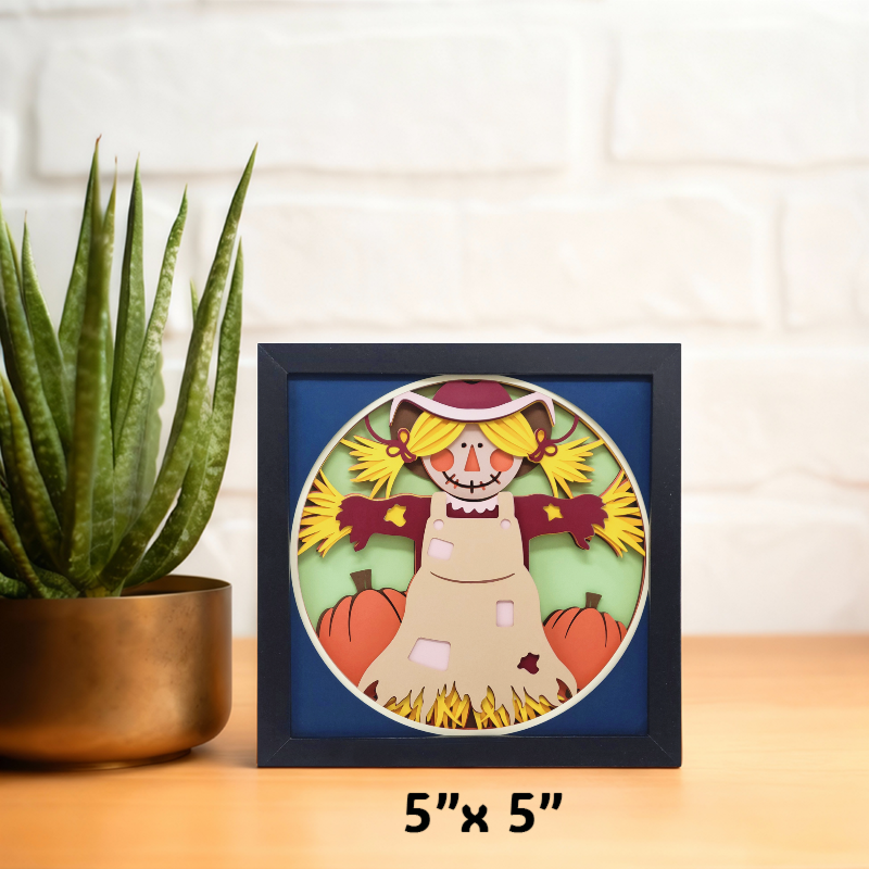 Framed artwork of a scarecrow with pumpkins on a table next to a plant.