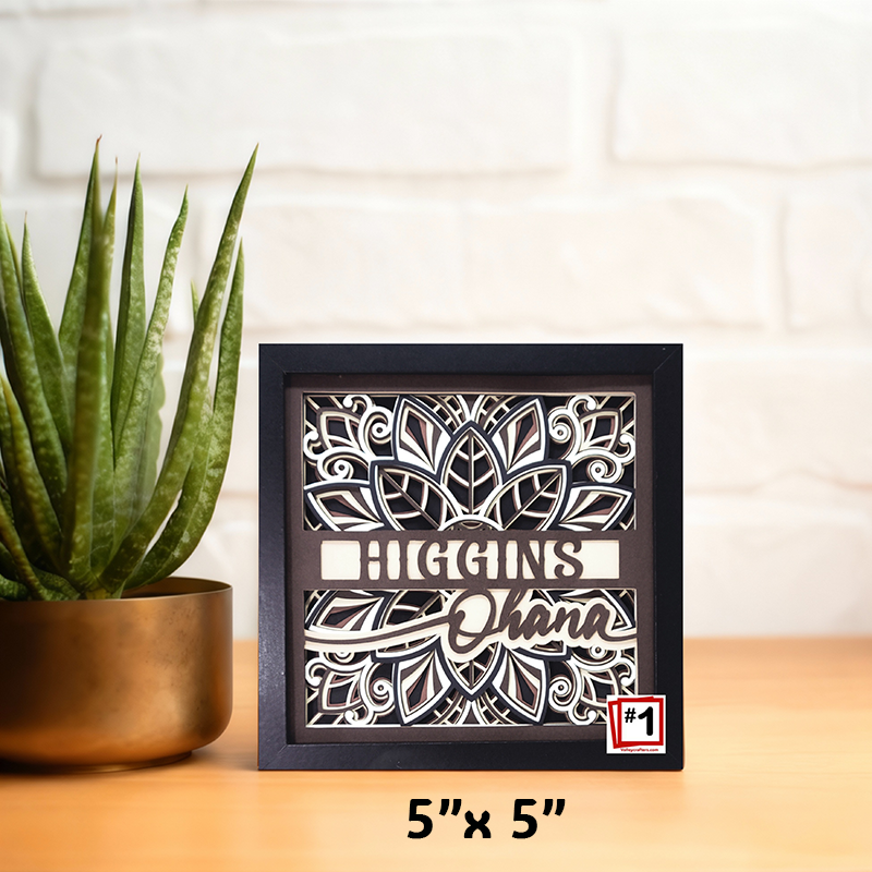 Decorative shadow box with cut-out design and text, placed on a surface with a plant in the background.