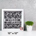 Framed decorative wall art with floral design and text on a gray surface with a plant.