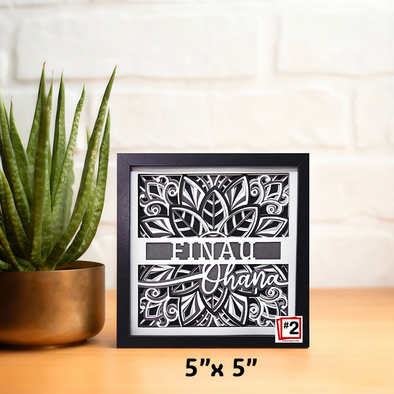 Framed decorative art piece with 'Finau Ohana' text and plant on a wooden surface