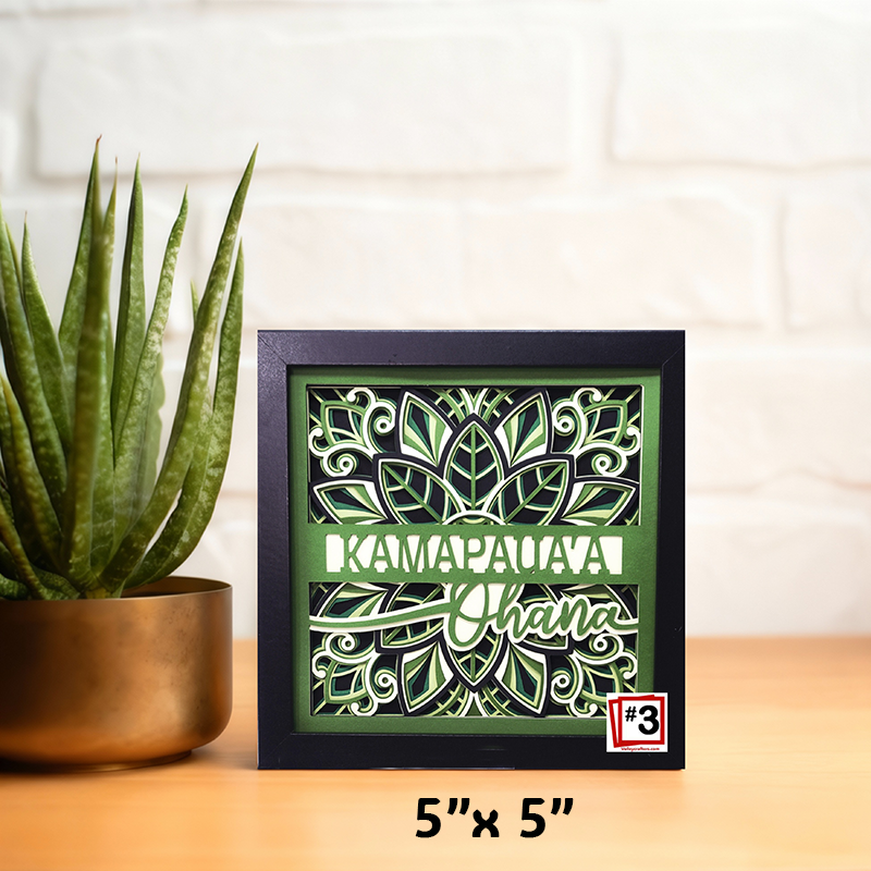 Decorative shadow box with green leaf pattern and text on a wooden surface with a plant.