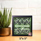 Decorative shadow box with green leaf pattern and text on a wooden surface with a plant.