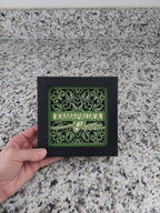 Framed decorative piece with green design held by a hand on a speckled surface
