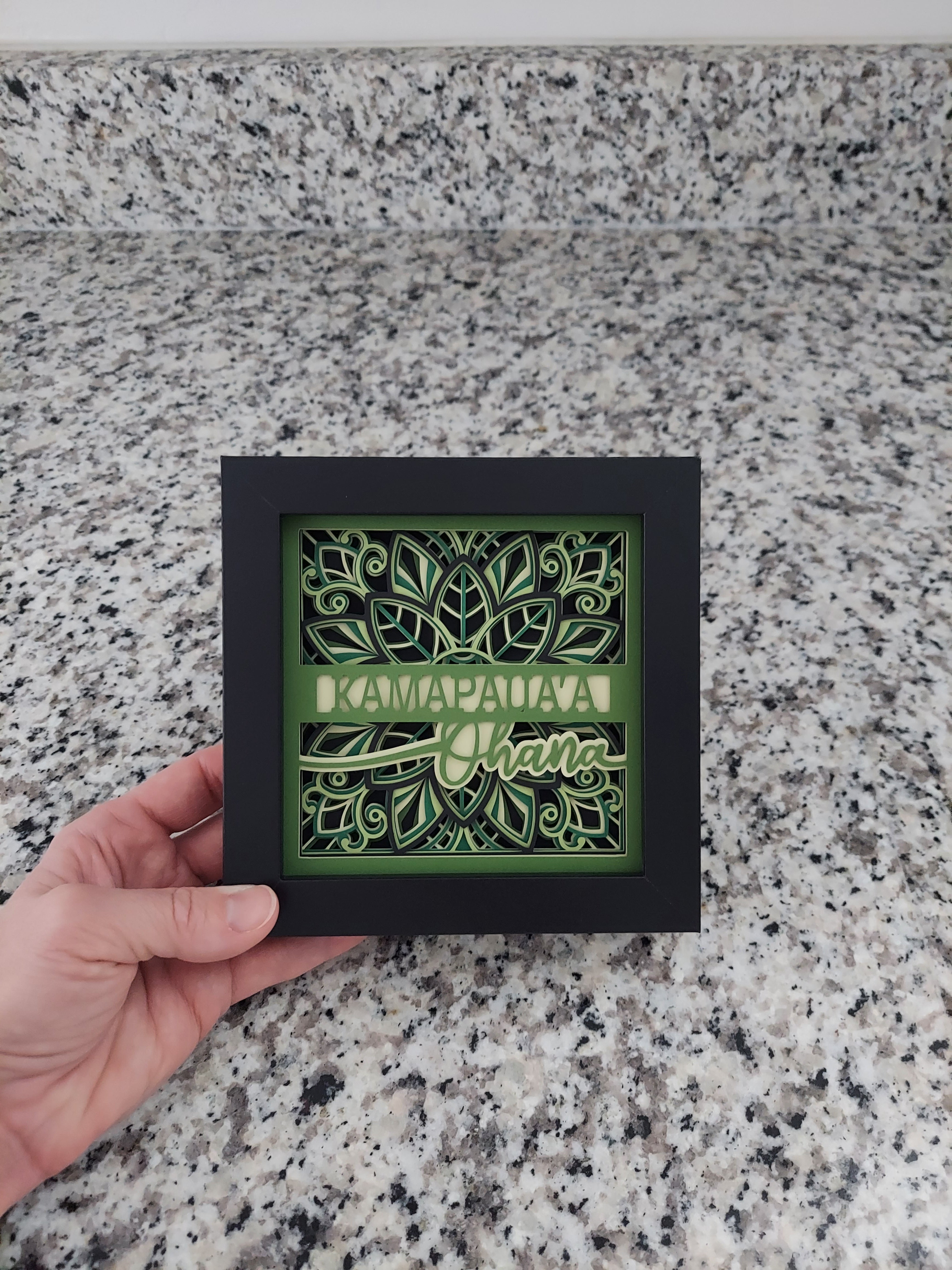 Framed decorative piece with green design held by a hand on a speckled surface