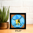 Framed art piece with a character holding a blue umbrella on a wooden surface with a plant.