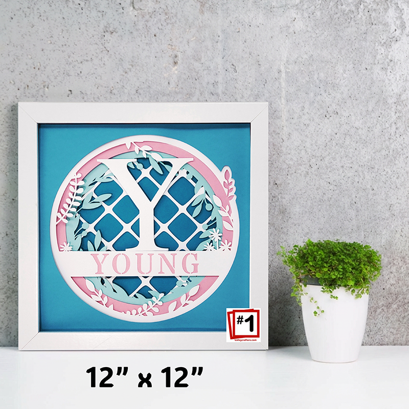 Framed artwork with 'YOUNG' text and decorative elements on a blue background, placed on a gray surface with a plant.