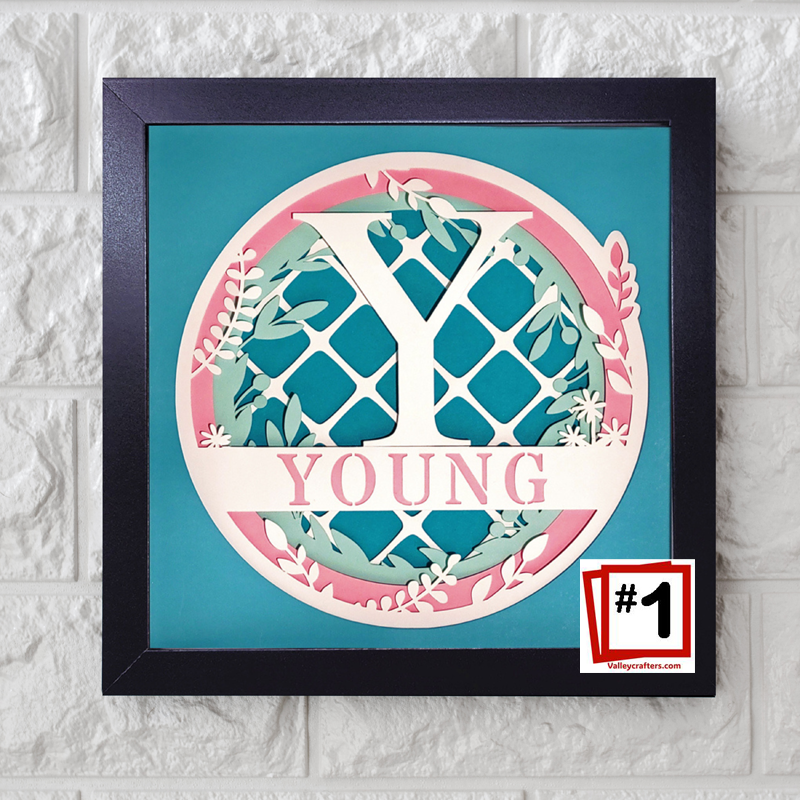 Framed decorative letter 'Y' with floral elements on a teal background, displayed on a textured white wall.