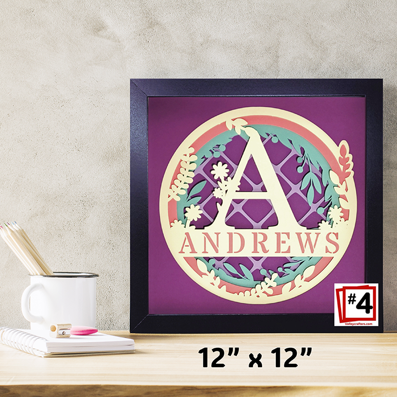 Framed decorative wall art with 'ANDREWS' and a floral design on a purple background, placed on a desk.
