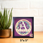 Decorative framed artwork with a monogram and 'ANDREWS' text, placed on a surface with a plant in the background.