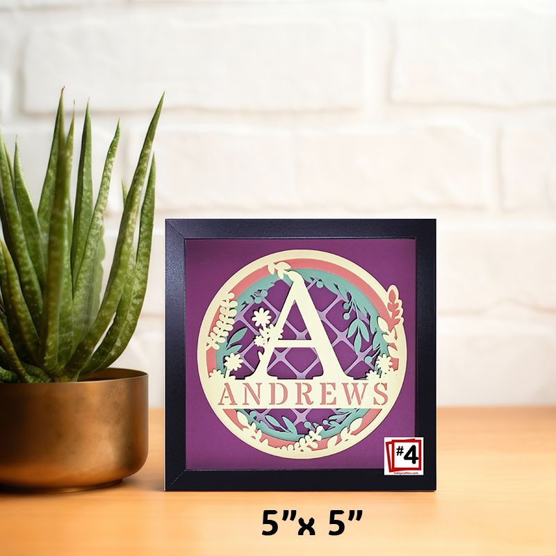Decorative framed artwork with a monogram and 'ANDREWS' text, placed on a surface with a plant in the background.