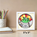 Decorative wall art with tulips and a watering can in a white frame, placed on a desk with stationery items.
