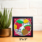 Small framed artwork with colorful fruit and vegetable shapes on a wooden surface with a plant in the background.