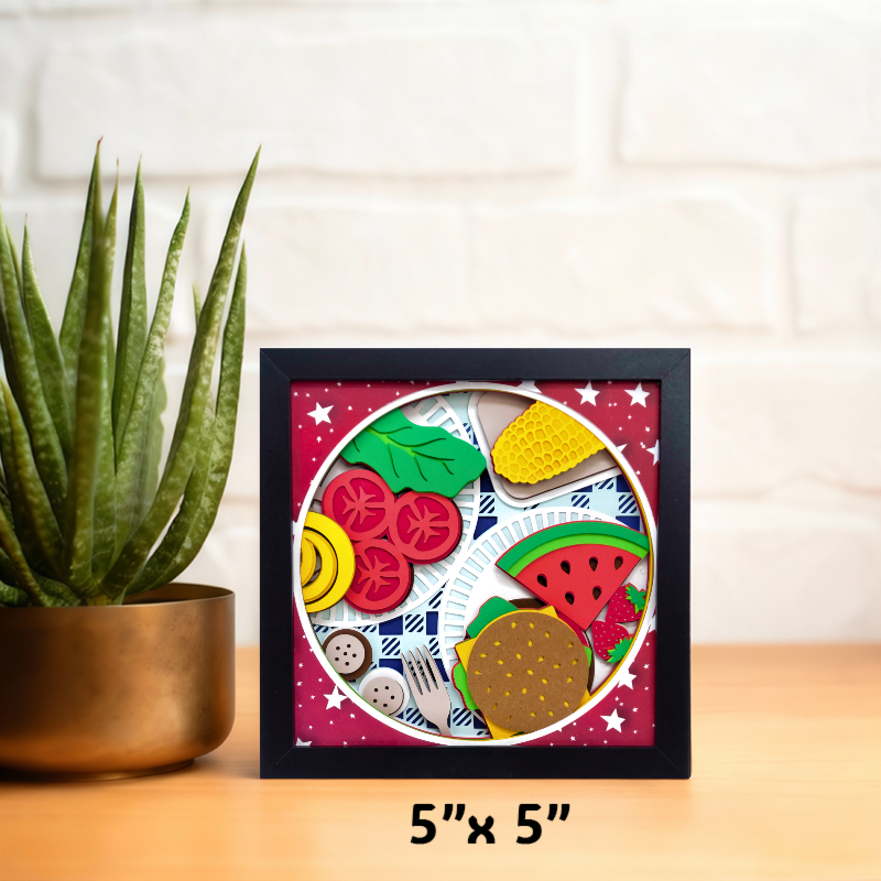 Small framed artwork with colorful fruit and vegetable shapes on a wooden surface with a plant in the background.