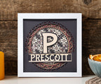 Decorative wall art with a stylized 'P' and 'Prescott' text in a white frame on a wooden surface.