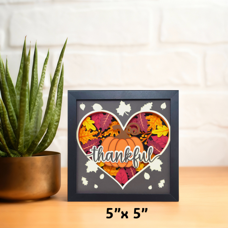 Decorative shadow box with autumn leaves and pumpkin design on a wooden surface with a plant.