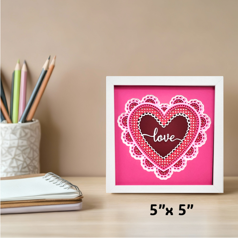 Framed heart-shaped artwork with 'love' text on a pink background, placed on a desk with stationery items.