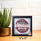 Framed decorative sign with 'the Blackburn family' text on a wooden surface with a plant.