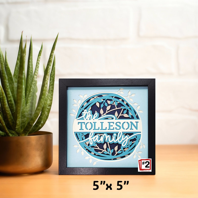 Framed decorative sign with 'the TOLLESON family' text, next to a potted plant on a wooden surface.