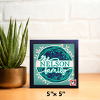Framed artwork with 'the NELSON family' text on a wooden surface with a plant.